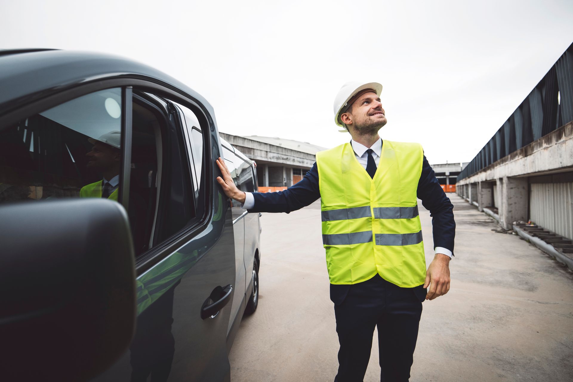 A construction worker in a yellow vest and hard hat stands next to a van, looking upwards in an industrial setting.