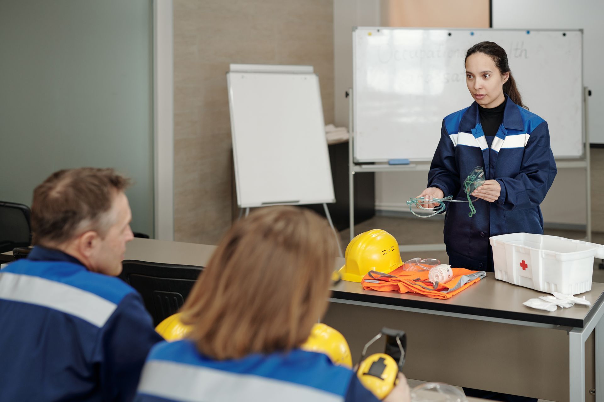 A woman in a blue uniform gives a presentation to a small group. She holds wires, and safety gear sits on a table.