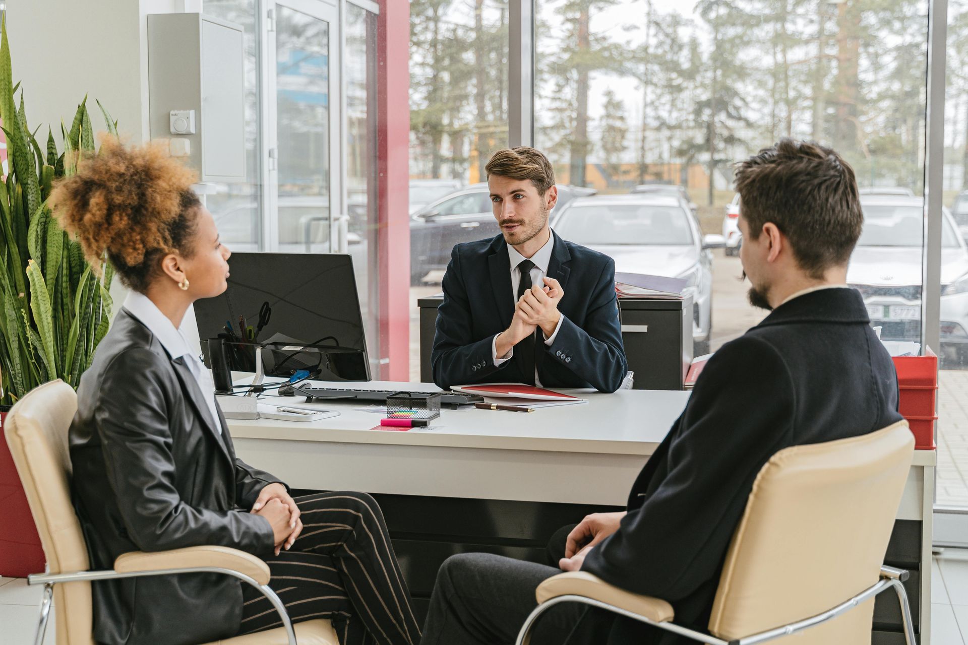 A car salesperson in a suit talks to a couple, discussing paperwork at a desk in a dealership.