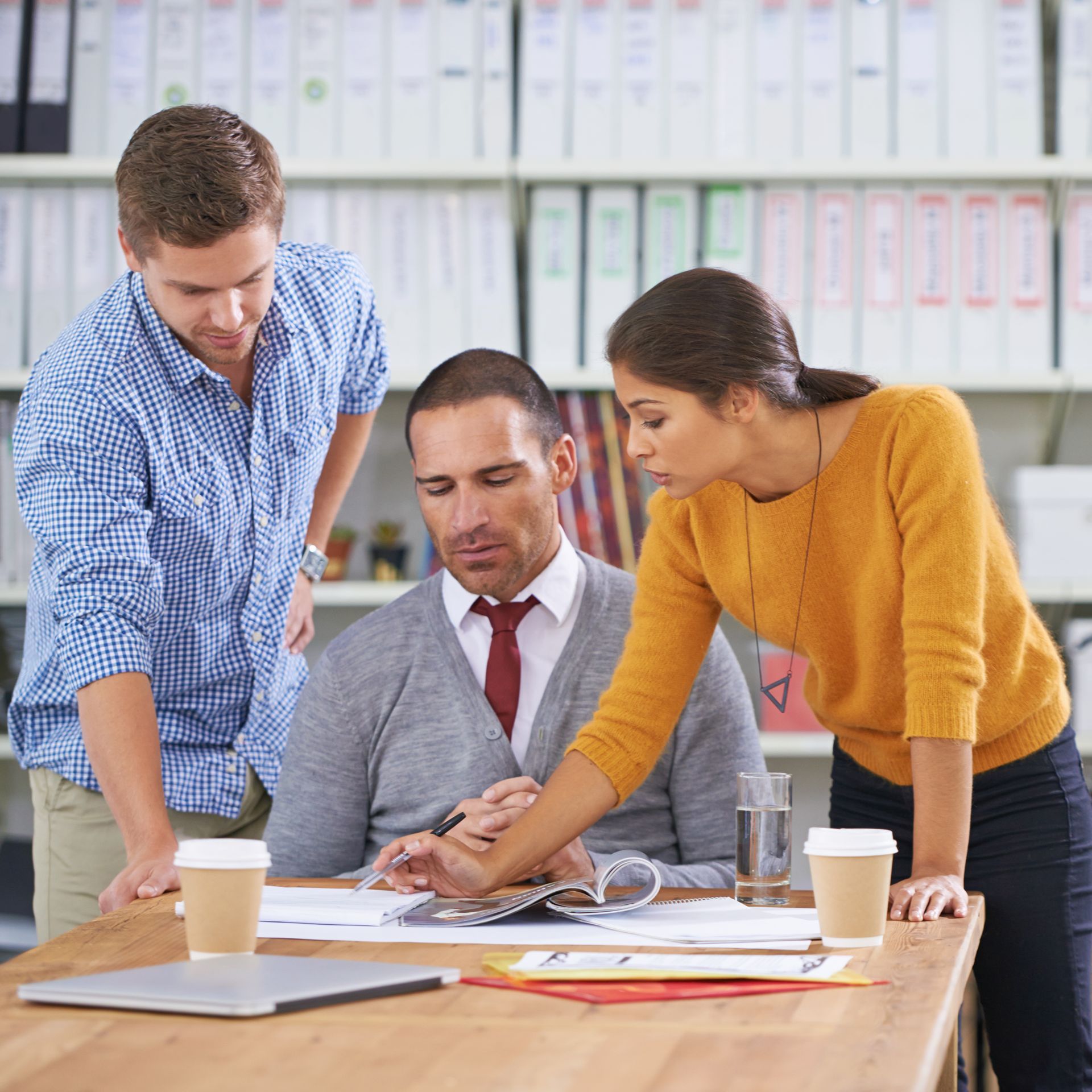 Three colleagues review paperwork at a desk in an office. A woman points to a document while two men look on.