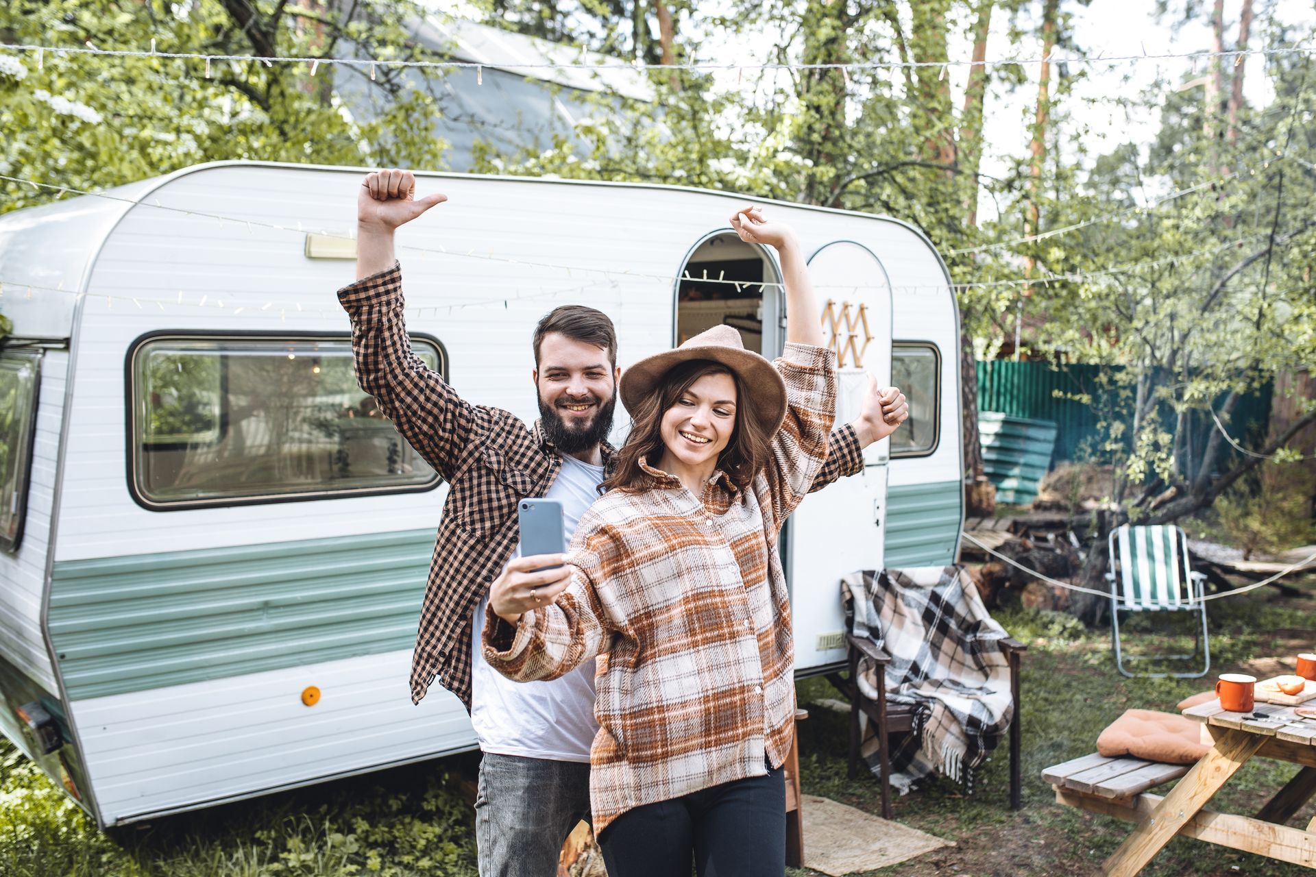 A man is playing a guitar in front of a camper van while a woman sits in chairs.