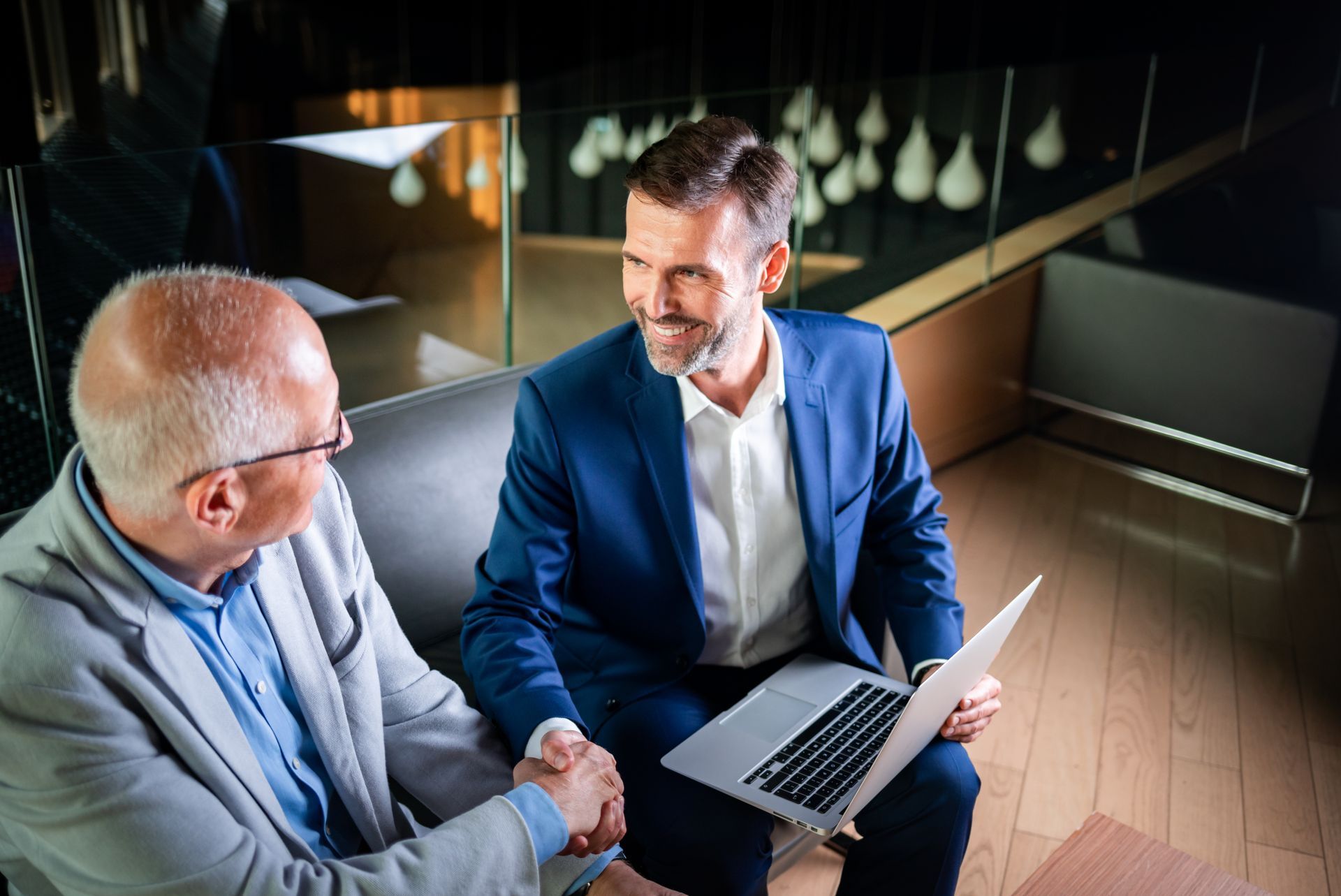 Two men in suits shaking hands, one holding a laptop, in a modern office setting.