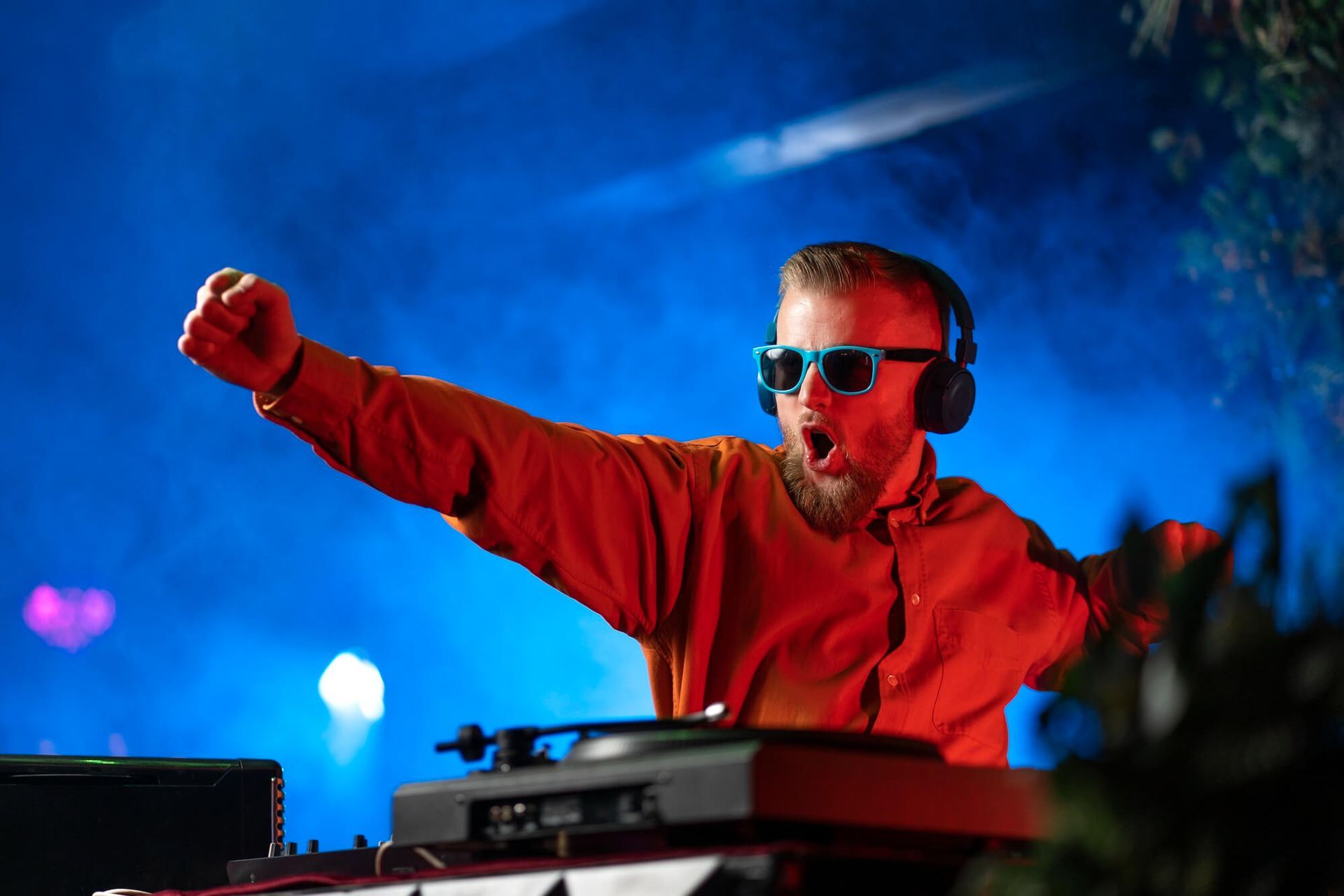 DJ with orange shirt, sunglasses, and headphones, fist raised, playing music at a blue-lit outdoor event.