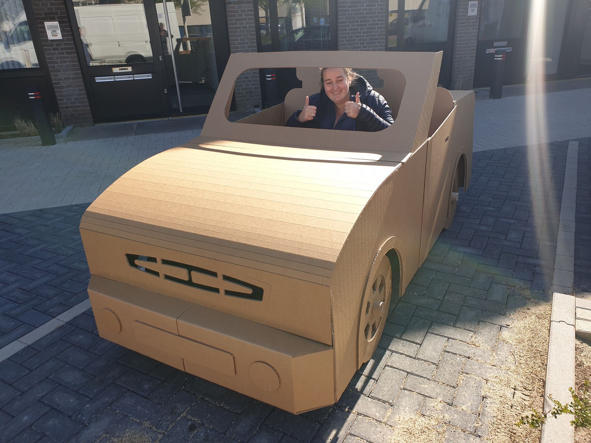 Een man zit in een kartonnen auto en steekt zijn duim omhoog