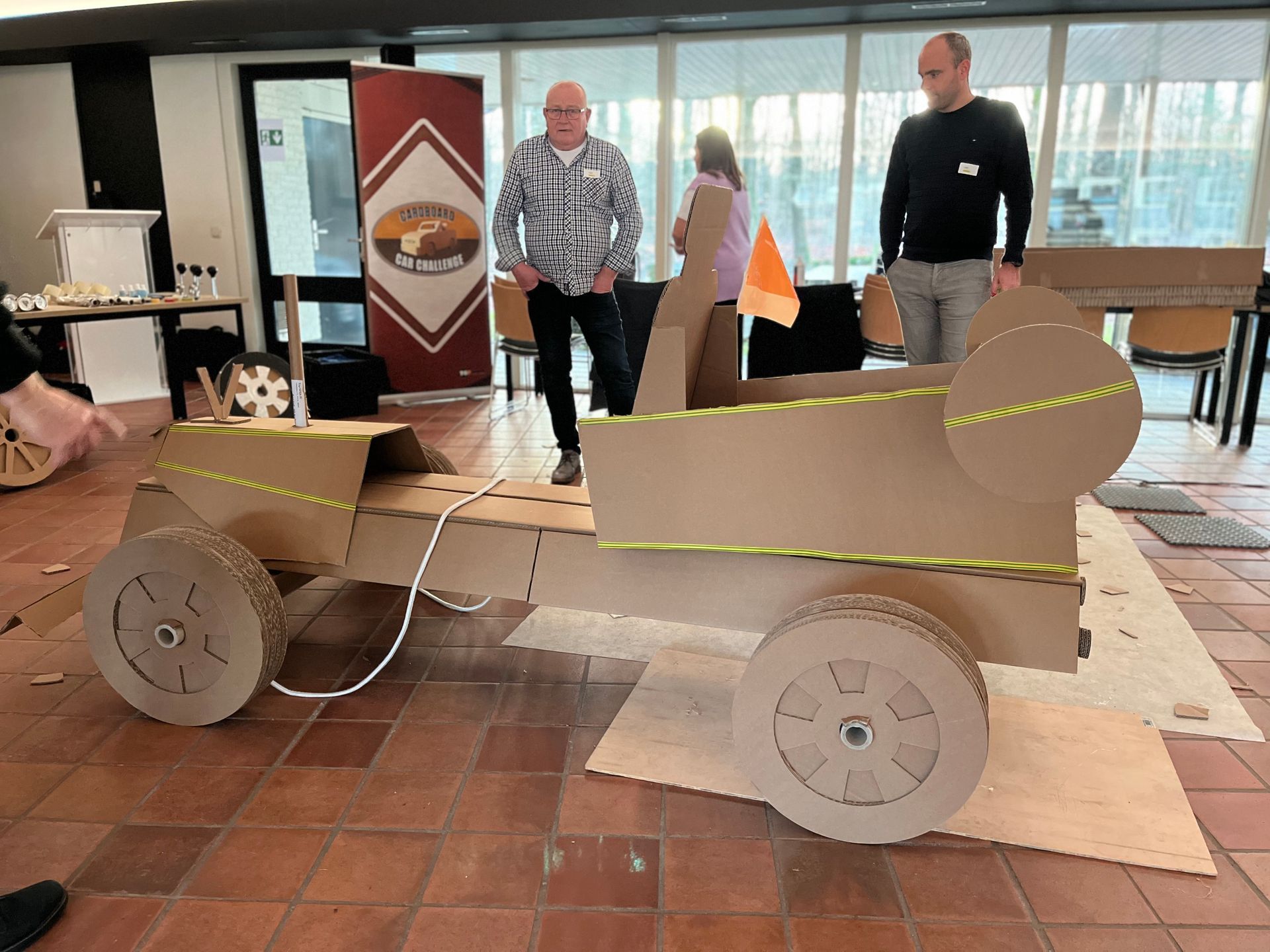 Twee mannen staan ​​naast een kartonnen auto op een tegelvloer.