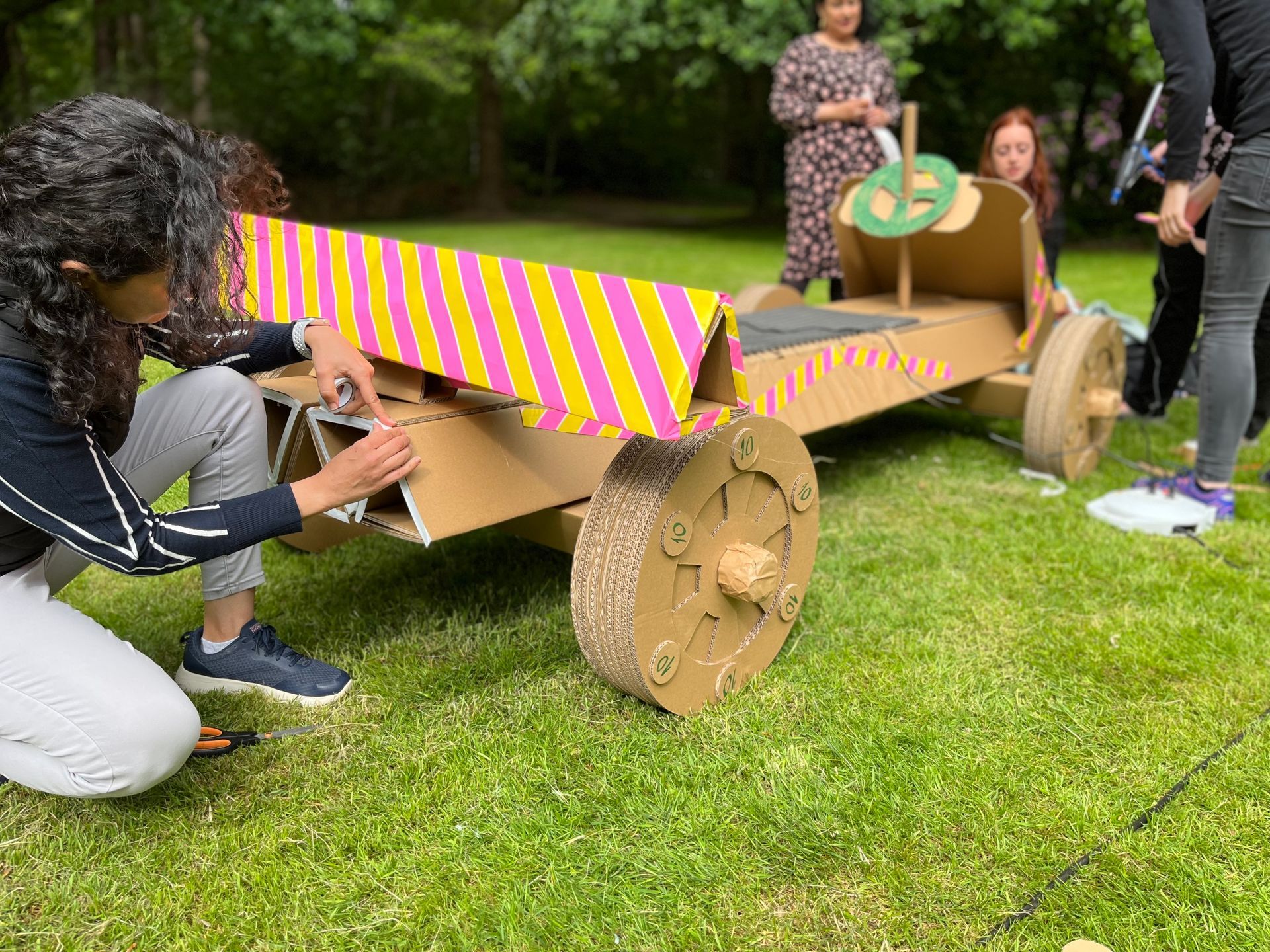 Een groep mensen werkt in het gras aan een kartonnen auto.