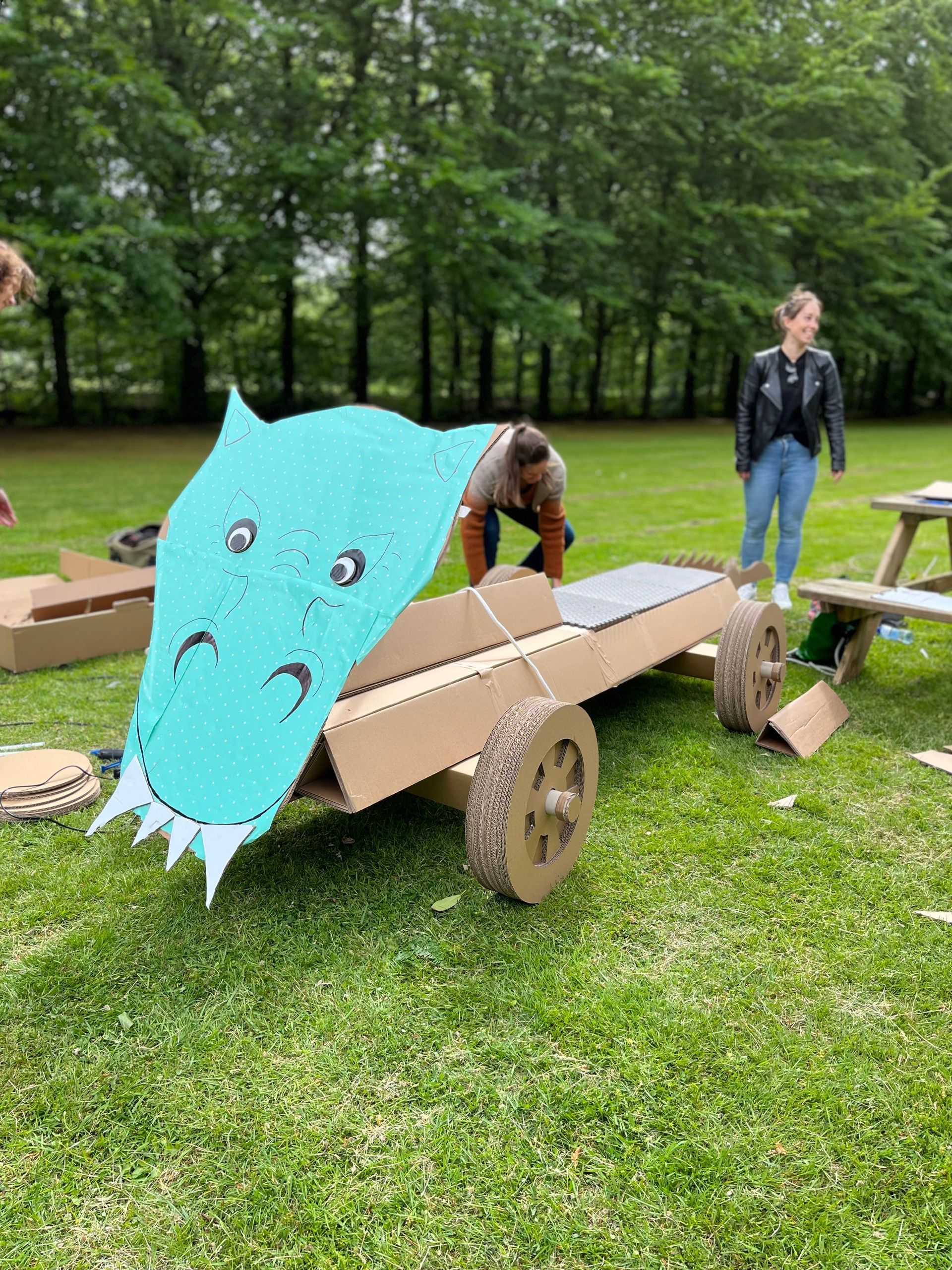 In een grasveld wordt een kartonnen auto gebouwd met een drakengezicht erop.