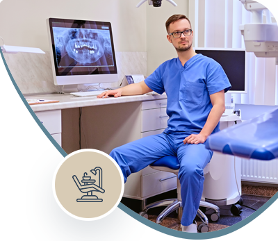 Dentist in blue scrubs seated beside a dental chair and computer in a bright clinic