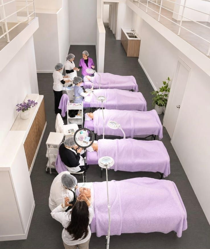 Sala de formación de un salón de belleza con personas practicando tratamientos en camillas cubiertas de tela morada.