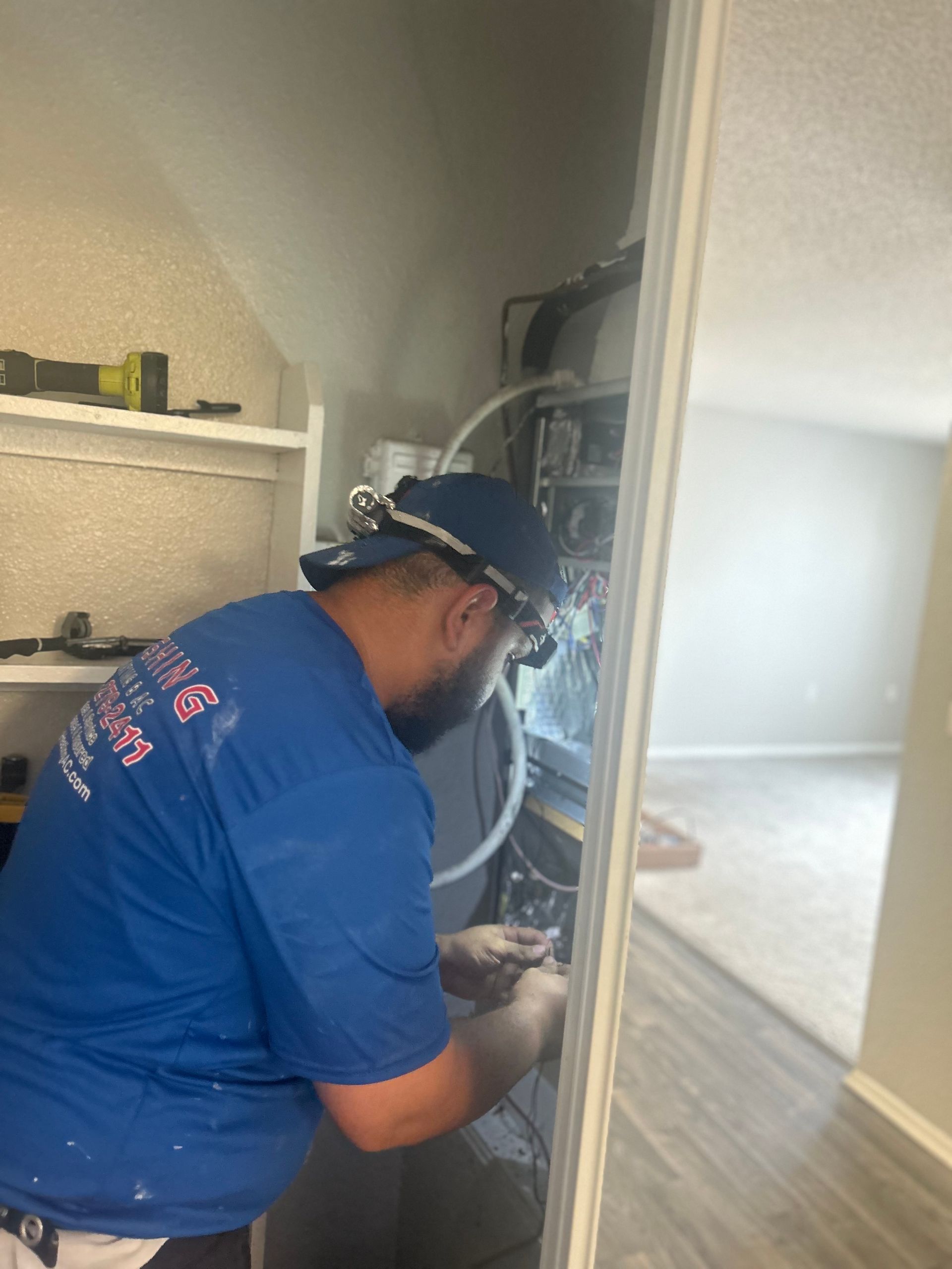 A person in a blue shirt with a headlamp working on a panel inside a utility closet.