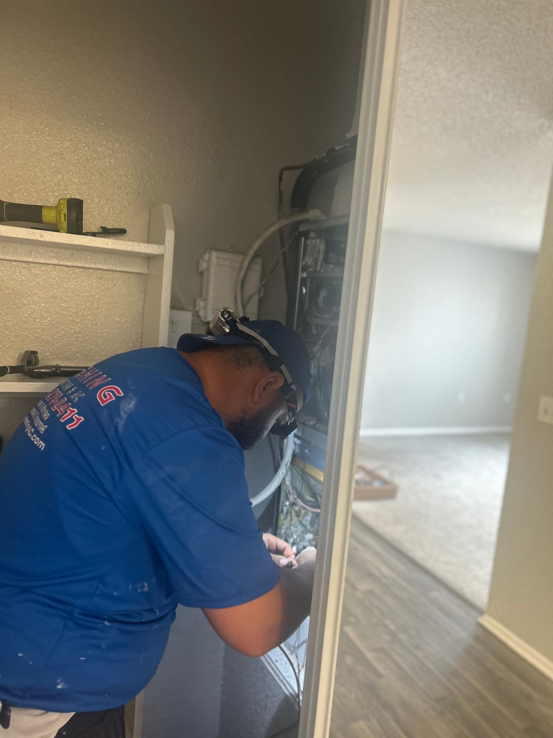 Man in blue shirt working on machinery in a closet. Bright light, white shelf, neutral-colored walls and carpet.