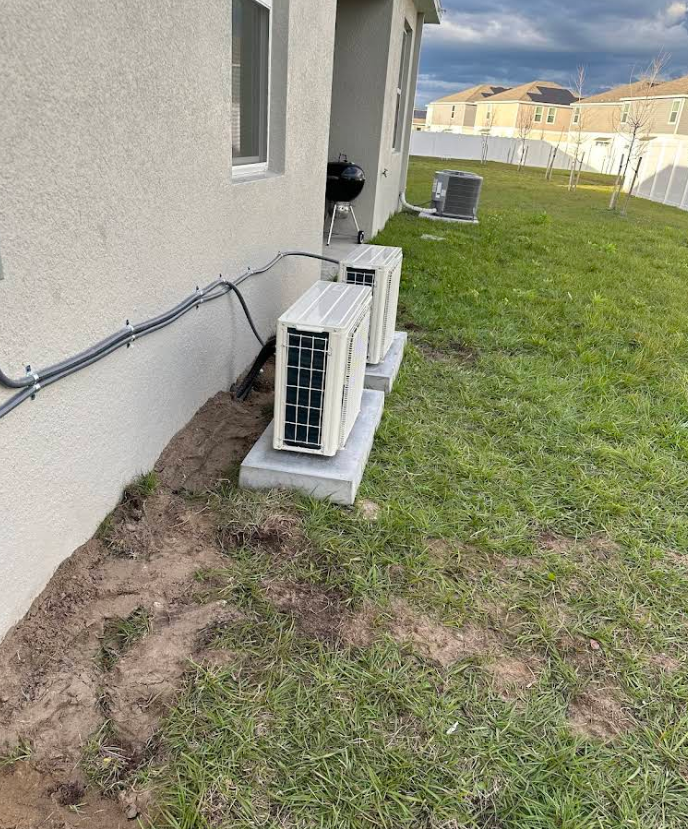 Two air conditioning units on concrete pads beside a light-colored building, outdoors on grass.