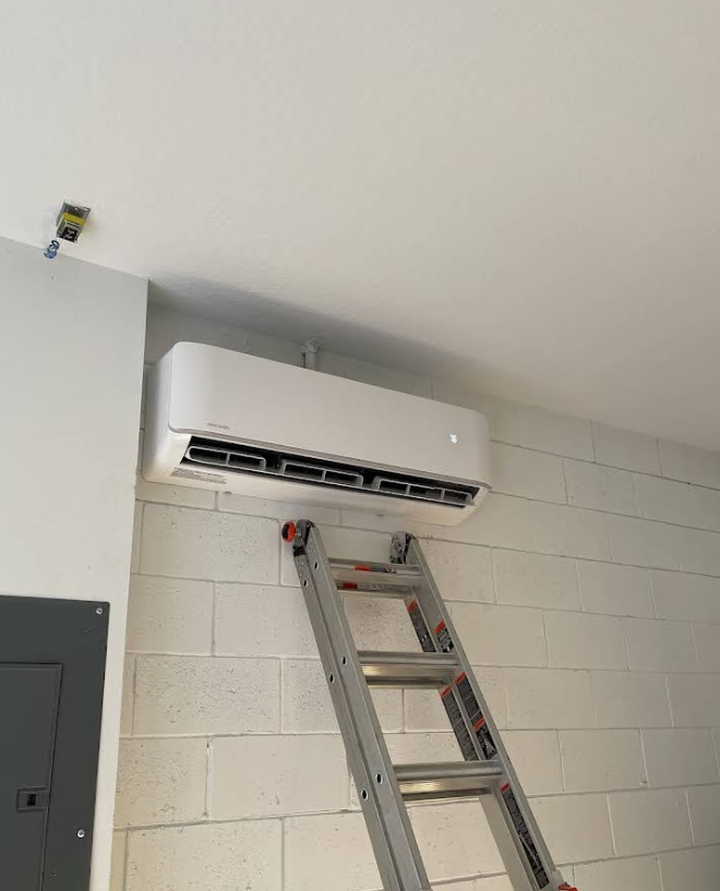 White air conditioning unit mounted on a white brick wall, with a ladder leaned beneath it.