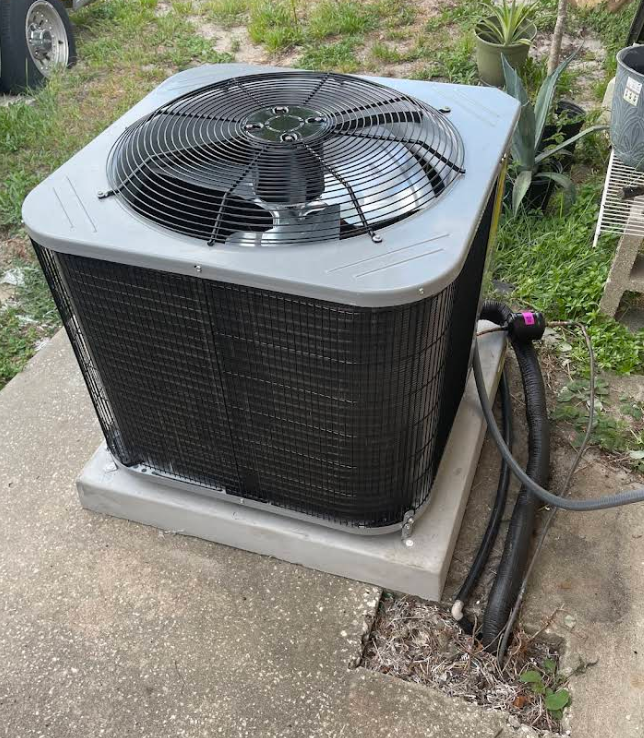 Outdoor air conditioning unit, gray and black, on concrete, with black pipes.