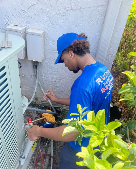 HVAC technician in blue shirt and cap working on an outdoor air conditioning unit.