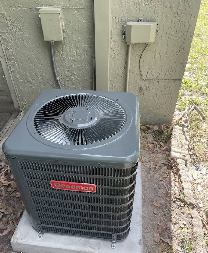 Goodman air conditioning unit outside a building, gray and red, with electrical boxes on the wall.