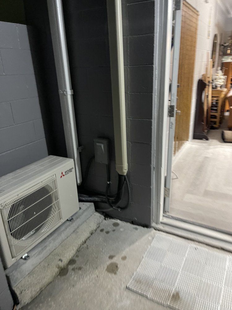 Outdoor air conditioning unit next to a wall with pipes and a doorway. Concrete floor.