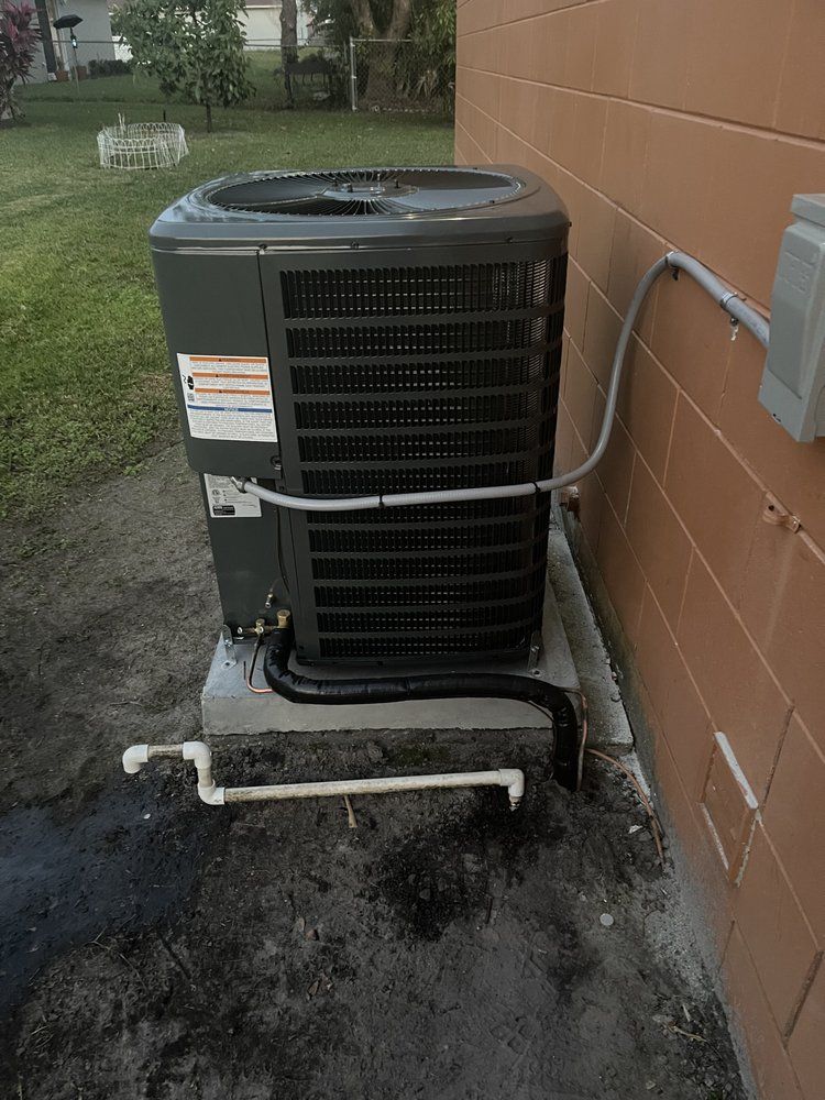 Gray air conditioning unit outside a brick building. Black pipes and a white drainage pipe visible.