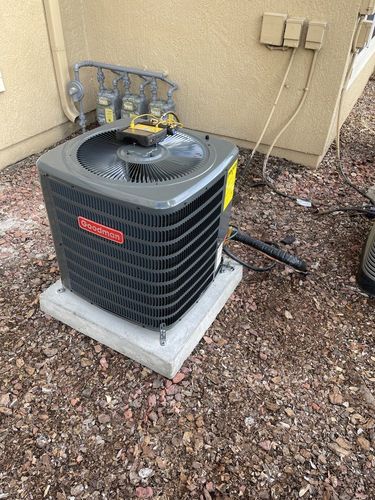 A Goodman air conditioner unit on a concrete pad, next to a beige building, set on mulch.
