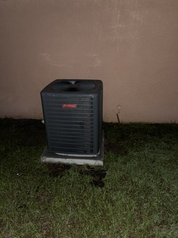 Air conditioning unit, dark gray, on a concrete pad, against a light brown wall, on grass.