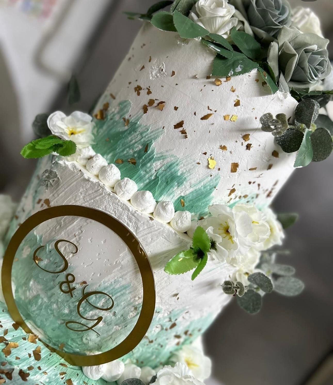 Torta bianca decorata con dettagli floreali verdi, puntini dorati e una scritta dorata 