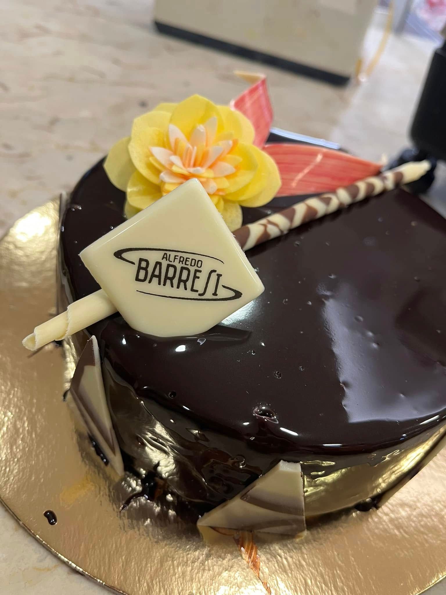 Torta al cioccolato con glassa lucida, decorazione con fiori gialli e targhetta BARRELI in cioccolato bianco sulla parte superiore.