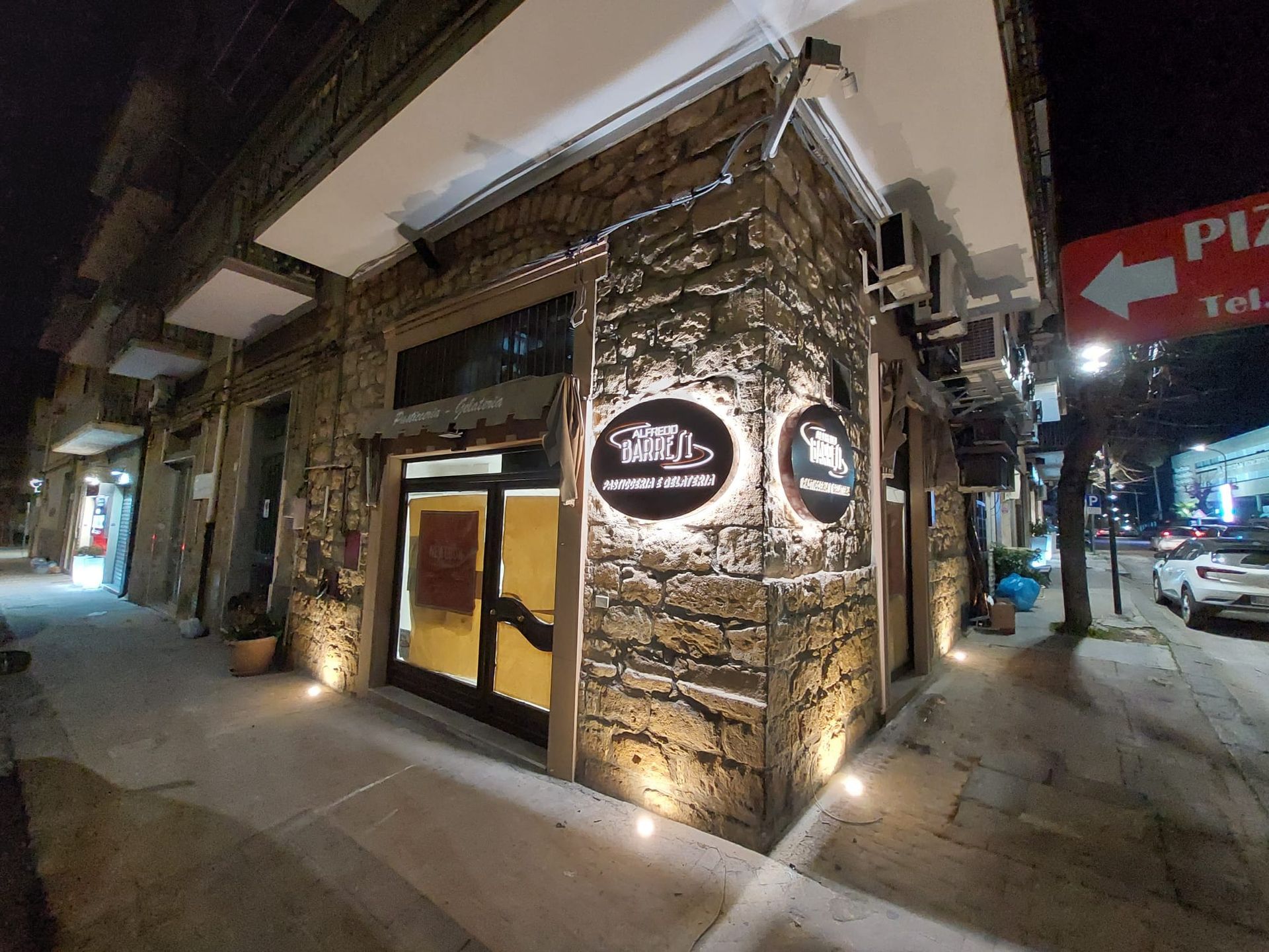 Angolo di strada notturno con un edificio in pietra illuminato e due insegne circolari luminose all'ingresso.