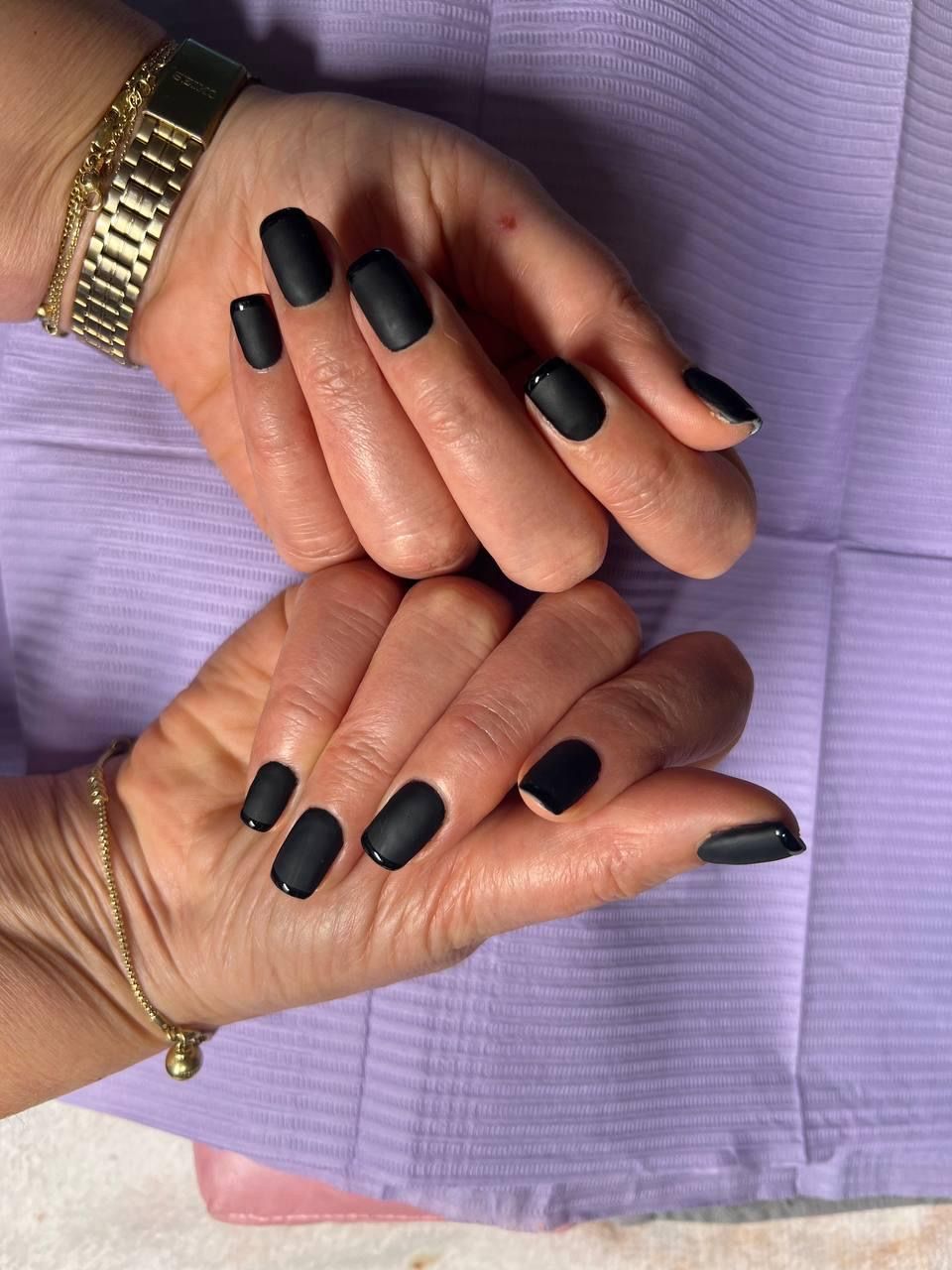 Hands with matte black nail polish, resting on a purple surface. A gold watch and bracelet are visible.