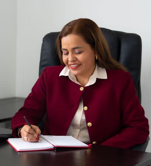 Una mujer con un blazer color burdeos sonríe mientras escribe en un cuaderno en un escritorio.