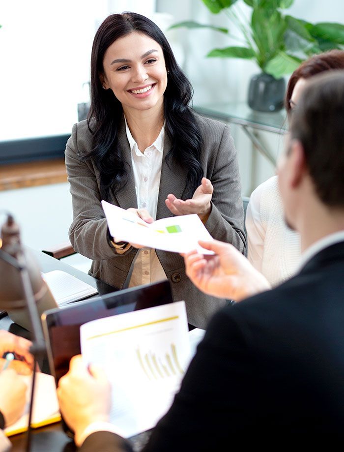Mujer con blazer pasando documentos a una persona, sonriendo en un entorno de oficina.