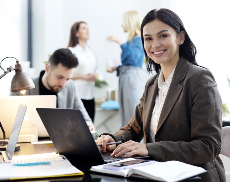 Una mujer con blazer sonríe mientras trabaja con una laptop en una oficina moderna. Compañeros al fondo.