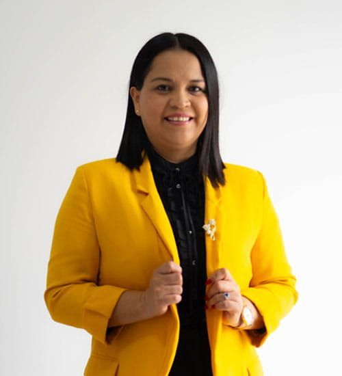 Mujer con blazer amarillo y camisa negra, sonriendo, con las manos cerca de la chaqueta sobre fondo blanco.