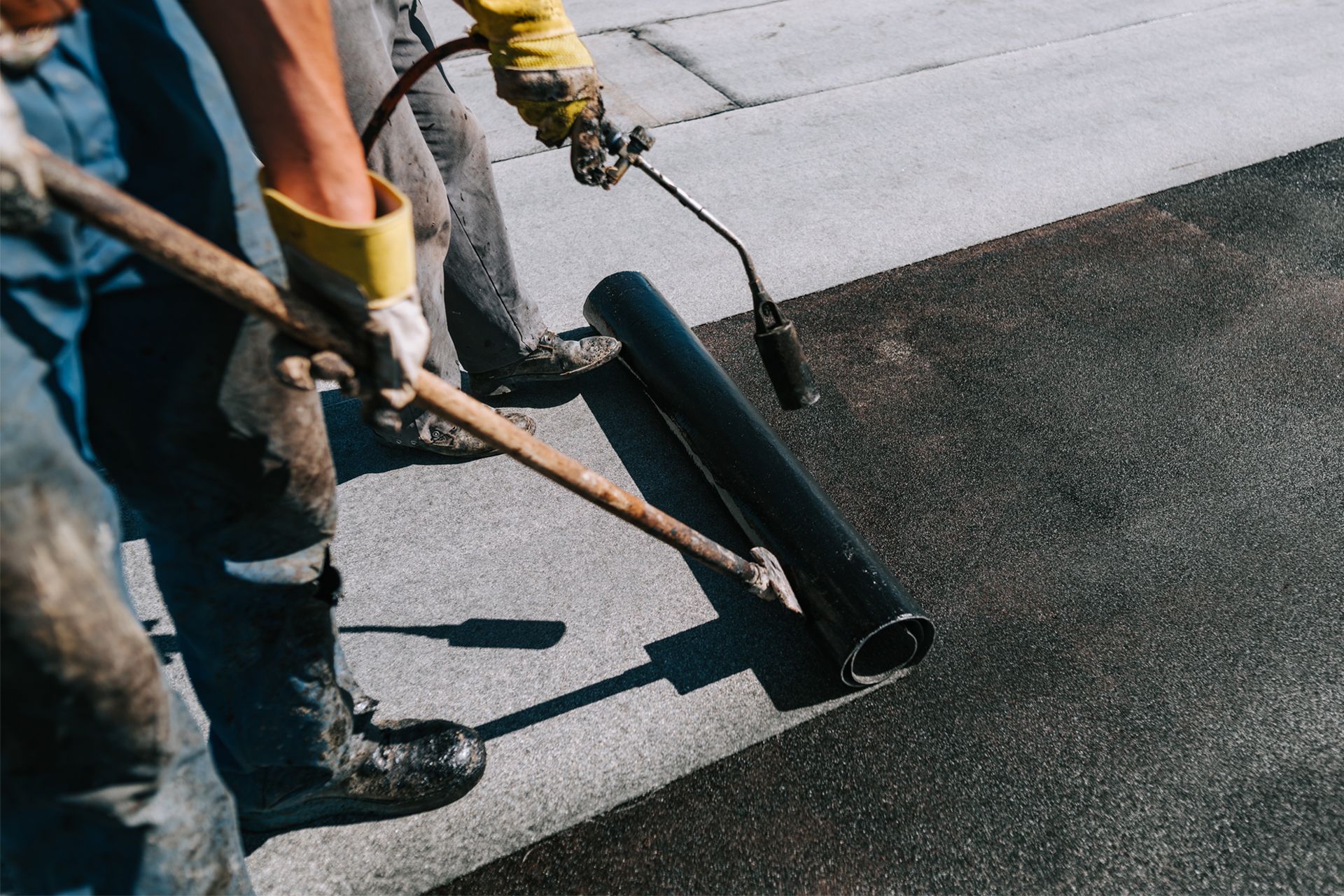 Two workers using a torch and roller to apply a dark, rolled roofing material on a flat roof.