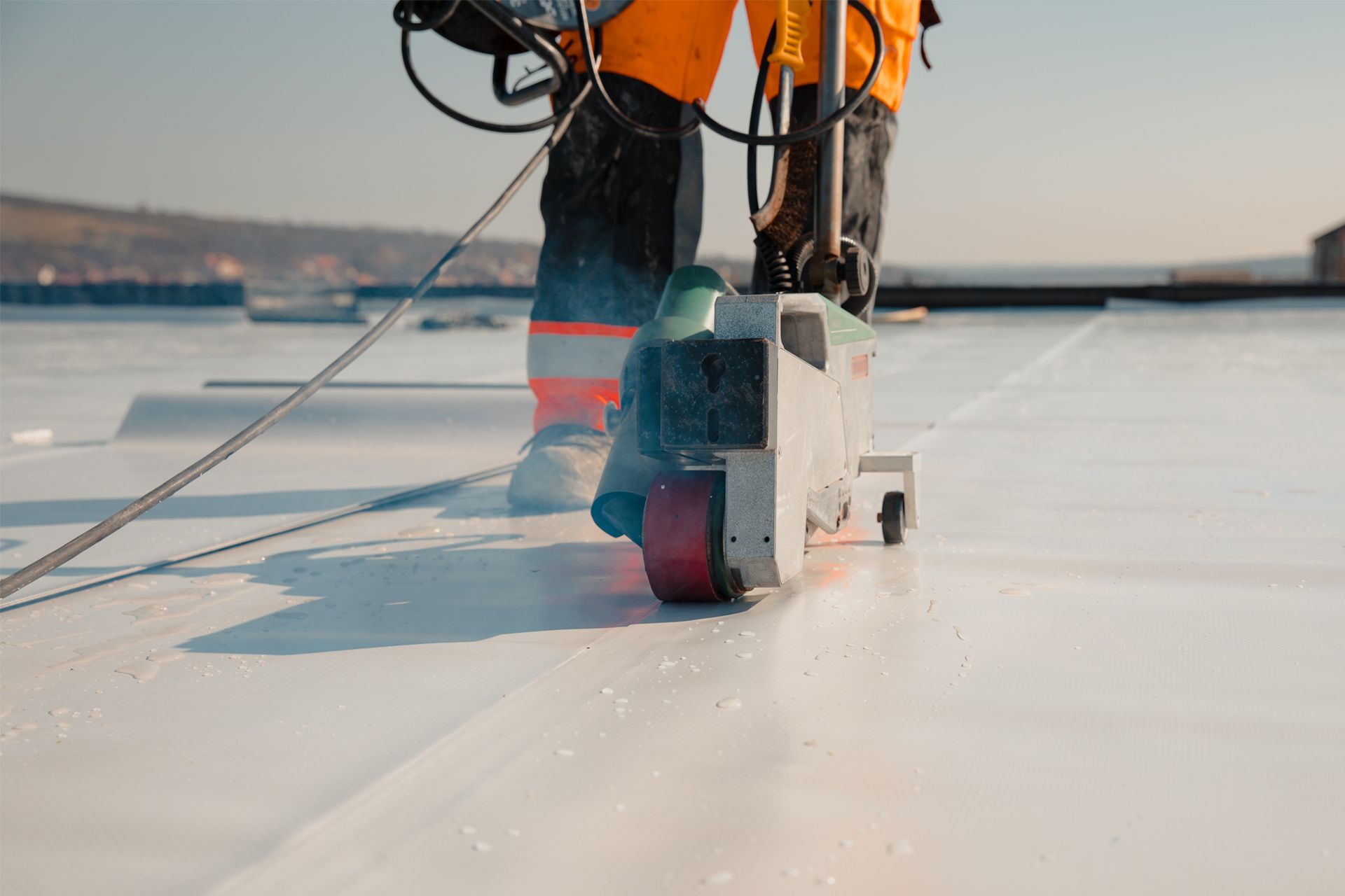 Roofer using a welding machine to seal white roofing material; worker in orange coveralls.
