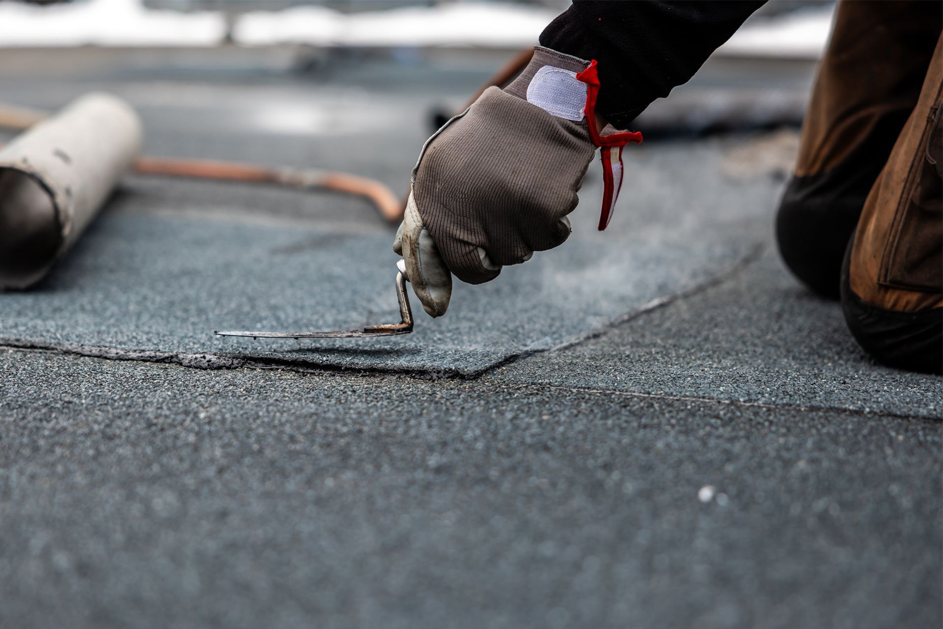 Roofer kneels, using a tool to repair a seam on a dark, textured flat roof.