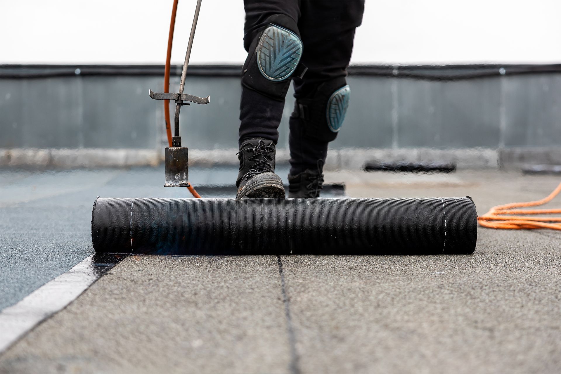 Roofer on a flat roof, rolling out roofing material, safety harness visible.