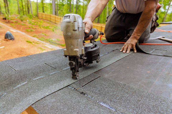 Roofer using a nail gun to install asphalt shingles on a roof, outdoors, sunny.