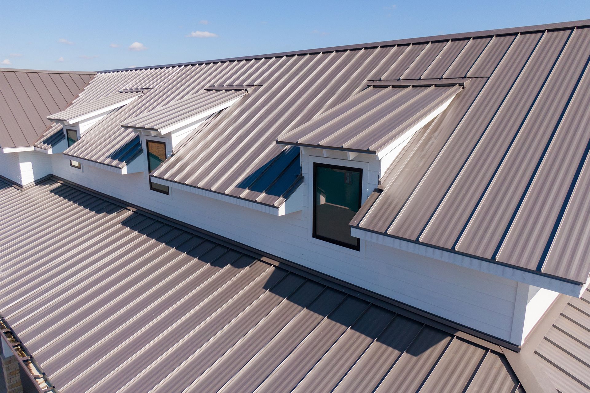 Gray metal roof with white dormers and windows against a blue sky.