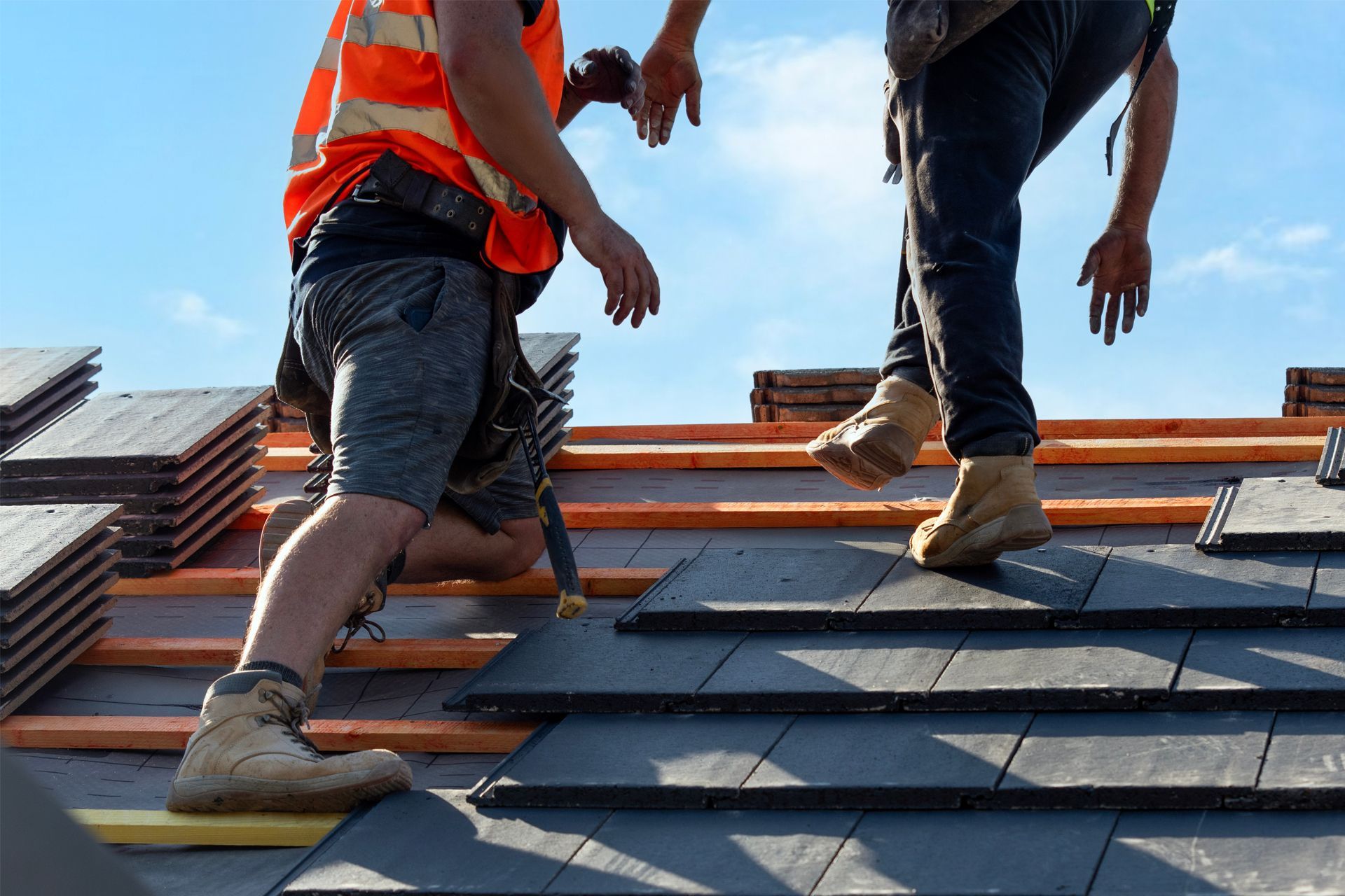 Two roofers installing dark gray tiles on a roof under a blue sky.