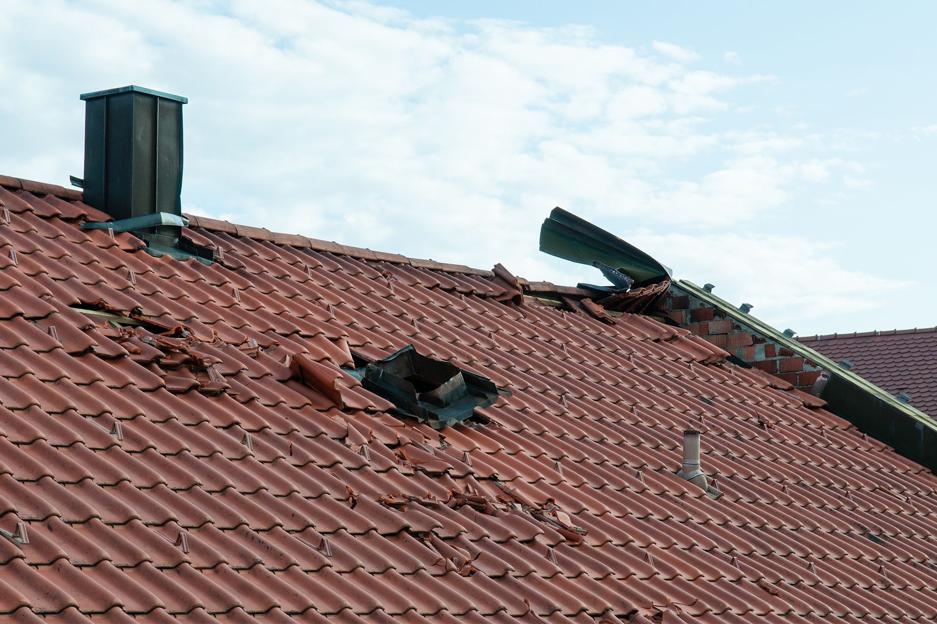 Red tiled roof with a damaged section, chimney, and gutter against a cloudy sky.