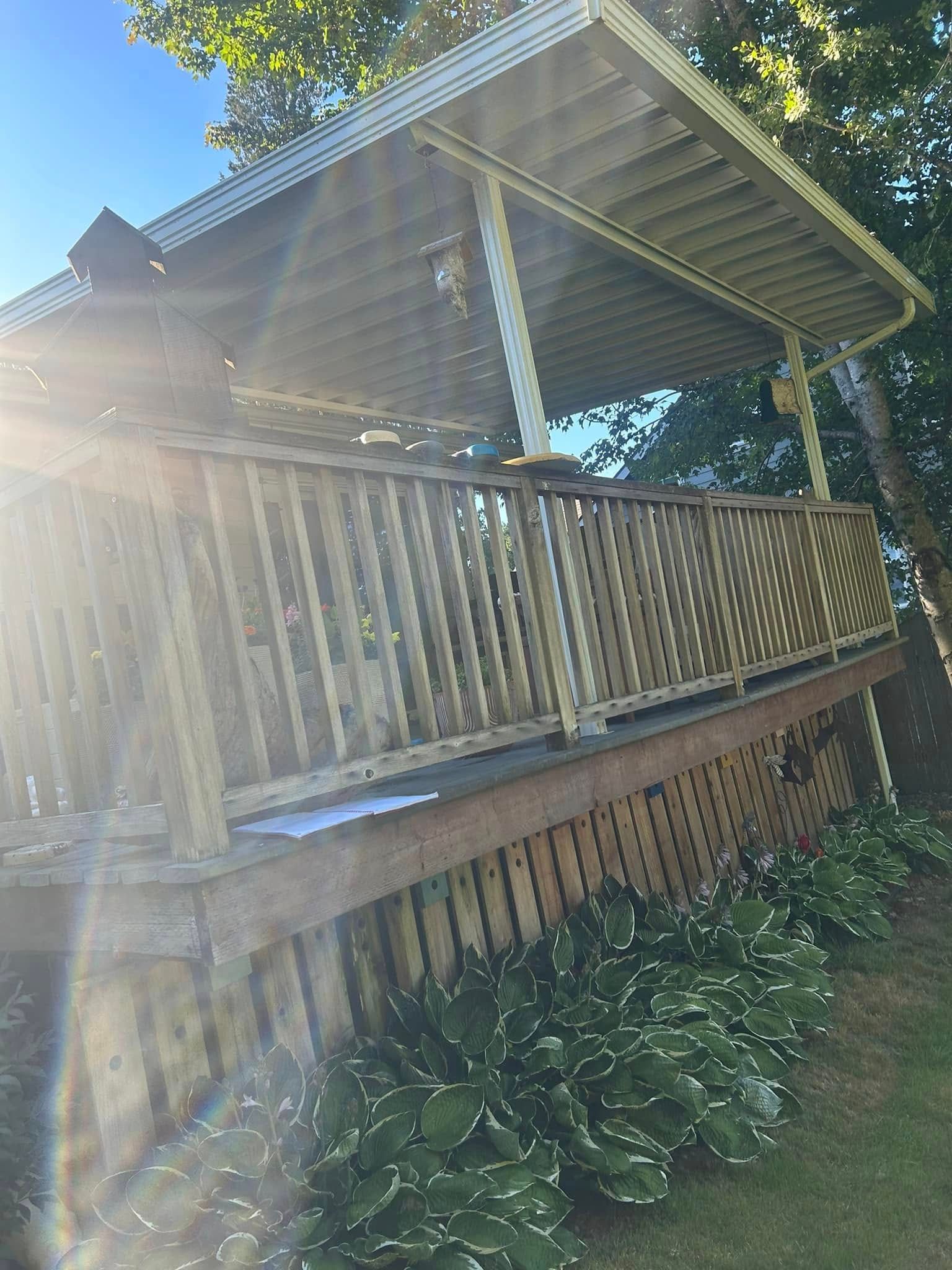 Wooden deck with railing, covered by a roof. Landscaping with green plants is below. Sunny day.