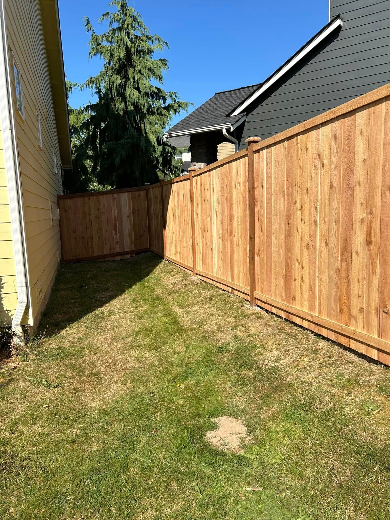 Wooden fence lines a small grassy backyard. A yellow house is on the left; a dark roof is above the fence.