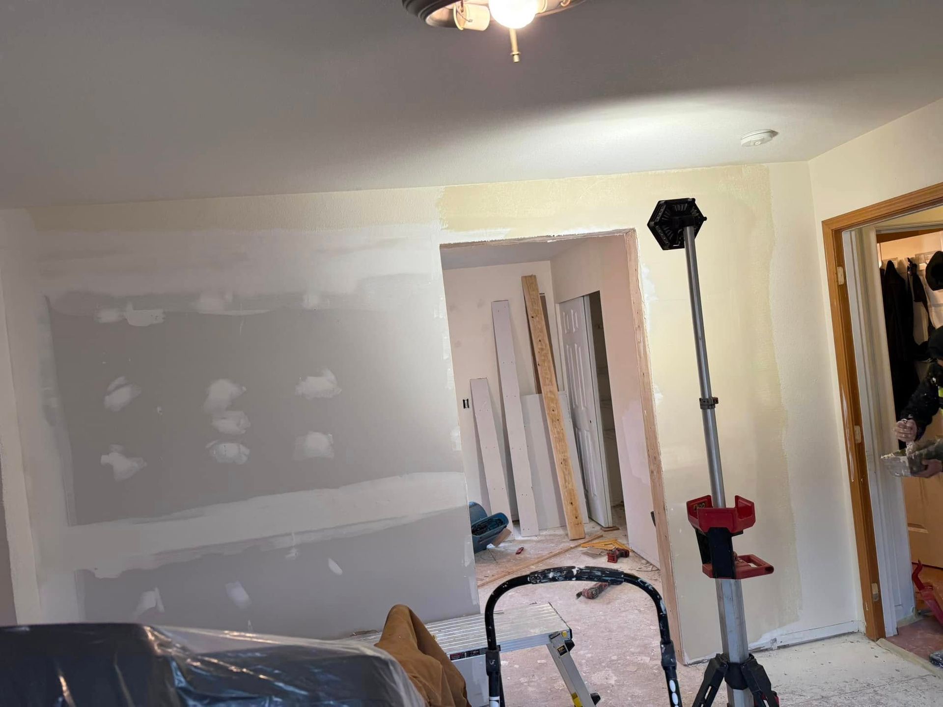 Room undergoing renovation, showing drywall, doorway, work light, and scattered materials.