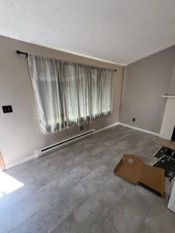 Empty living room with gray walls, flooring, and sheer curtains. Cardboard box and fireplace visible.