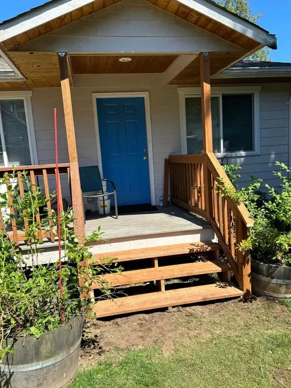Wooden porch with steps leading to a blue door. Beige house with wooden trim and railing.