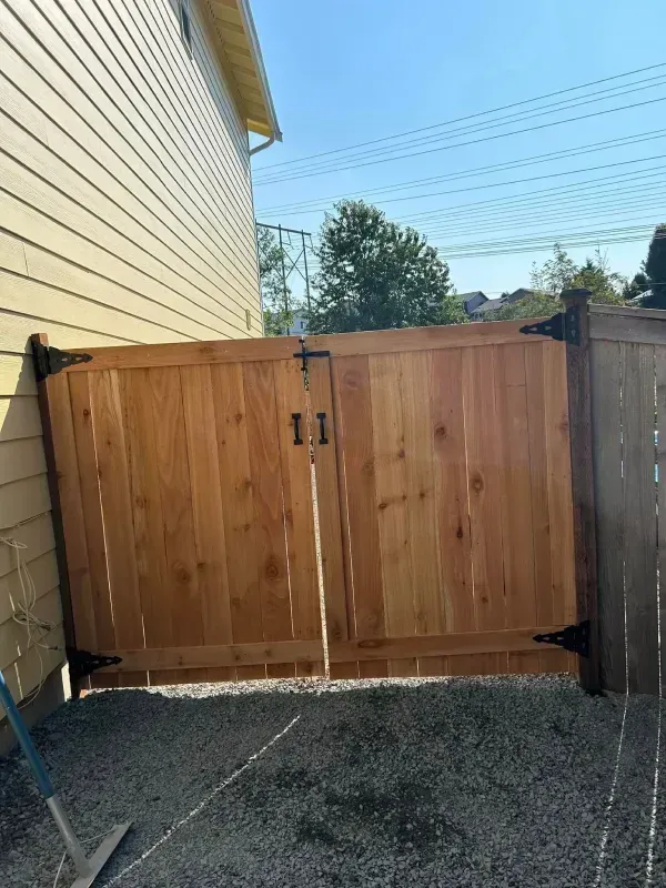 Wooden double gate in a gravel yard, attached to a tan building and a wooden fence; blue sky.