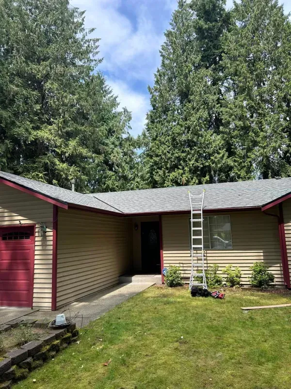 A tan house with a gray roof and a red door and trim. A ladder is leaning against the roof.