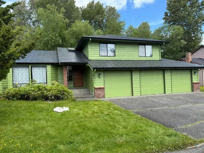 Green two-story house with a three-car garage, gray driveway, and lush green lawn in front.