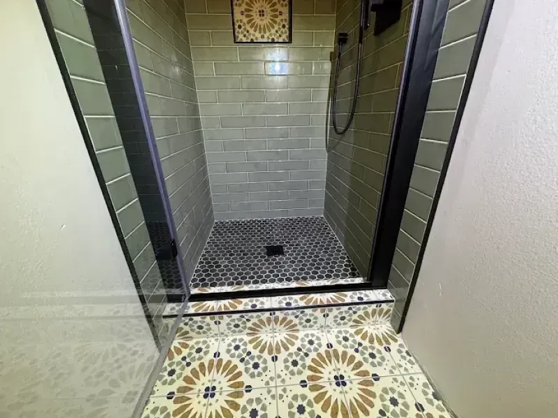 Shower with green tile walls, black-framed glass door, decorative floor tile outside, dark floor inside.