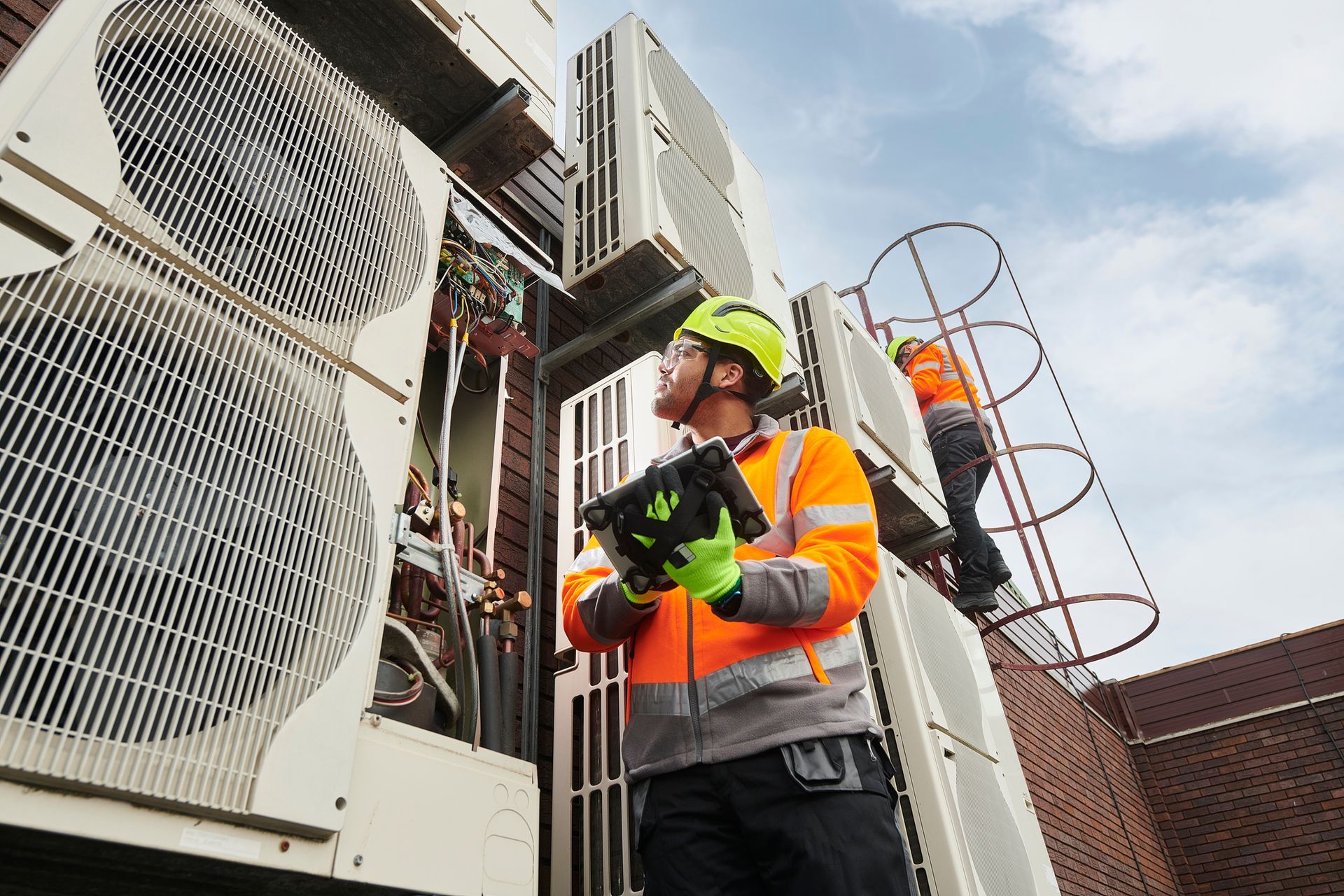 Technicians servicing large HVAC units on wall, one climbing ladder, both in safety gear.